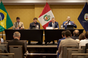 Fotografia de uma mesa composta por integrantes do poder judiciário. Quatro homens vestidos formalmente . Ao fundo parede de tom amarelo e bandeiras do Brasil, Peru e do poder Judiciário.