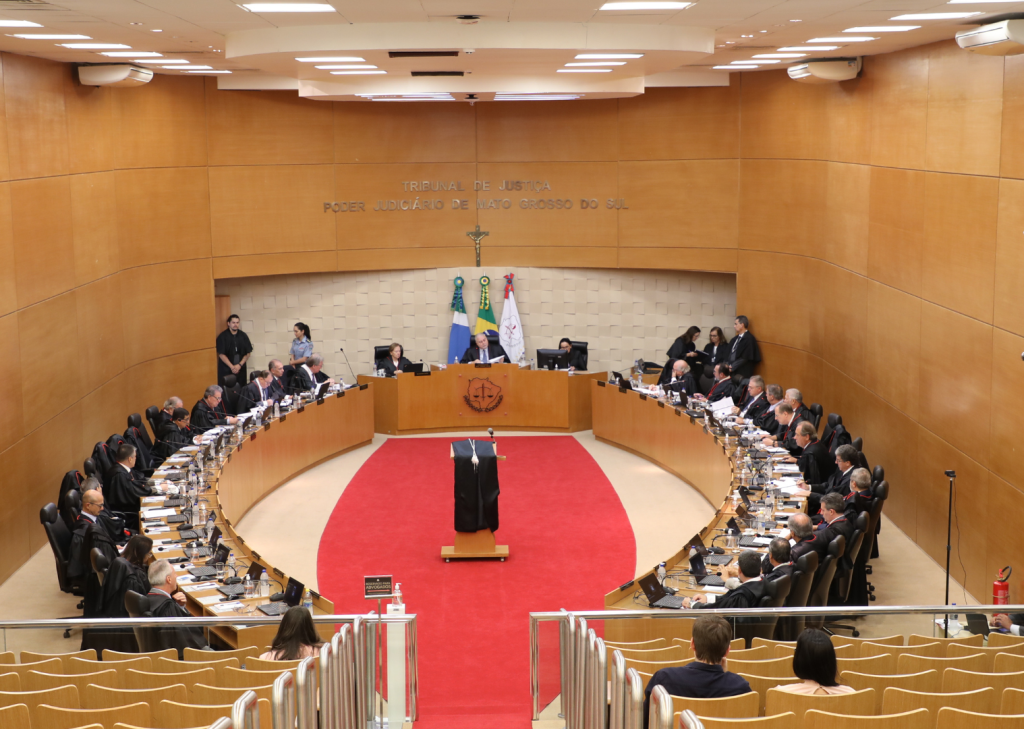 Fotografia em plano geral: Mesa plenária do Tribunal de Justiça do Mato Grosso do Sul, em formato circular de tom amadeirado, composta por autoridades do poder judiciário. A sala possui paredes de tom amadeirado, ao centro da mesa, tapete de tom vermelho. Ao fundo o letreiro: TRIBUNAL DE JUSTIÇA - PODER JUDICIÁRIO DE MATO GROSSO DO SUL.