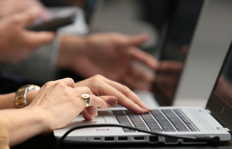Fotografia das mãos de uma mulher enquanto digita no teclado de um computador. No fundo desfocado a outra mão de um homem manuseando seu computador.