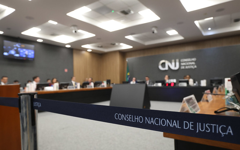 Fotografia do plenário do Conselho Nacional de Justiça (CNJ). Ao fundo desfocado, há pessoas sentadas em frente a monitores. Ao centro e em destaque, uma faixa com o texto: Conselho Nacional de Justiça.