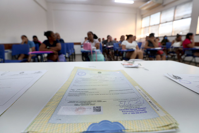 Fotografia de uma sala, com um documento em destaque no centro da imagem. No fundo, há um desfoque nas pessoas sentadas.