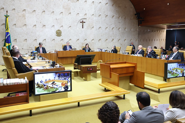 Fotografia dos ministros do Supremo Tribunal Federal (STF) em destaque no centro, além de telas reproduzindo a imagem deles. Acima dos ministros há um crucifixo e um brasão do Brasil. No canto esquerdo, uma bandeira do Brasil. No canto inferior direito, pessoas acompanham a sessão plenária.