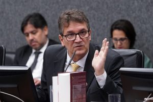 Fotografia do Conselheiro do CNJ Alexandre Teixeira, na 13ª Sessão Ordinária, enquanto fala em um microfone. Ele usa um terno com uma gravata amarela e óculos. Há também um livro com capa vermelha na frente do conselheiro. 