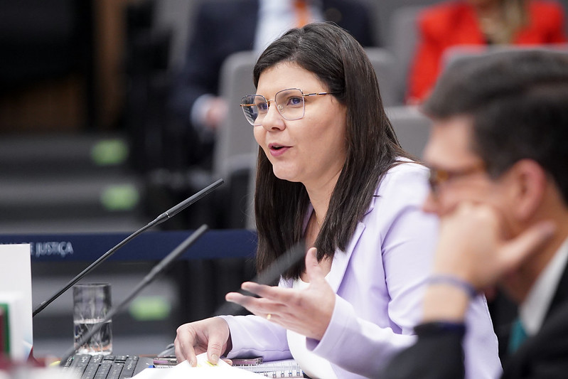 Fotografia da Conselheira do CNJ Daiane Nogueira de Lira, na 13ª Sessão Ordinária, enquanto fala em um microfone. Ela usa um blazer roxo e óculos. 