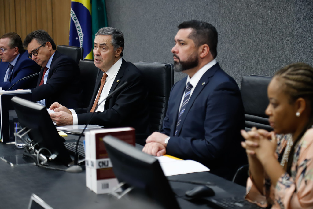 Fotografia da 13ª Sessão Ordinária onde o presidente do Conselho Nacional de Justiça (CNJ) e do Supremo Tribunal Federal (STF), Ministro Luís Roberto Barroso (ele usa um terno preto com gravata laranja) fala em um microfone que está a sua frente. Do lado esquerdo da foto está o Dr. Mansour Elias Karmouche da Ordem dos Advogados do Brasil (de terno e gravata em tons de azul) e o Dr. José Adonis Callou de Sá da Procuradoria Geral da República (que usa terno preto e gravata vermelha). Do lado direito da foto, está o Presidente dos Correios Fabiano Silva dos Santos (de terno e gravata em tons de azul) a Dra. Adriana Cruz, Secretária Geral do CNJ (que usa uma blusa em tons de laranja claro e flores azuis e brancas estampadas). 