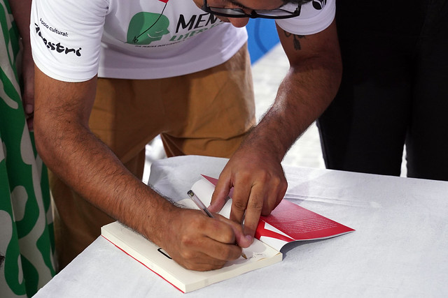 Fotografia de um homem assinando um livro de capa vermelha que está em cima de uma mesa. Ele veste uma blusa branca com a inscrição “Mentes Literárias – da Magia dos Livros à Arte da Escrita” e uma calça bege. Ele também usa óculos de armação preta. 