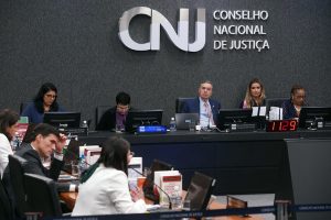 Fotografia da 14ª Sessão Ordinária de 2024 do CNJ. Da esquerda para a direita: Da esquerda para a direita: Dra. Claudia Lopes Medeiros, Conselheira Federal da OAB (de blusa azul e óculos de armação preta); Dra. Paula Bajer, da Procuradoria-Geral da República (de blusa roxa e blazer preto e usa óculos de armação transparente); Presidente do STF e do CNJ, Ministro Luís Roberto Barroso (que veste uma camisa social azul, uma gravata vermelha e um terno azul); e Dra. Adriana Cruz, Secretária-Geral do CNJ (que veste uma blusa azul, brincos e um colar prata). No canto inferior esquerdo alguns conselheiros do CNJ estão sentados, porém de costas. Em frente ao Ministro Barroso, há um relógio que marca 10:00 e ao fundo o logotipo do Conselho Nacional de Justiça (CNJ) na parede, juntamente com a inscrição “Conselho Nacional de Justiça”.