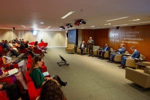 Fotografia de um auditório onde diversas pessoas, sentadas em poltronas vermelhas, assistem a um homem de terno cinza que está em pé e fala em um microfone. Outros quatro homens (três de terno azul e um terno cinza) sentam-se em poltronas ao redor do homem que está de pé.  Ao fundo, atrás dos homens, a logotipo e a inscrição da “Escola Nacional de Formação e Aperfeiçoamento de Magistrados”. 