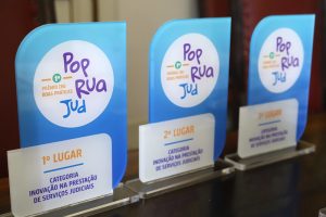Fotografia de três estatuetas do Prêmio PopRuaJud. Todas elas possuem a mesma composição. De fundo azul com uma inscrição nas cores roxa, azul e amarela, apresenta o texto: “1º Prêmio CNJ Boas Práticas” e “PopRuaJud”. Cada prêmio possui também uma inscrição. Da esquerda para direita: “1º Lugar Categoria Inovação na Prestação de Serviços Judiciais”; “2º Lugar Categoria Inovação na Prestação de Serviços Judiciais”; e “3º Lugar Categoria Inovação na Prestação de Serviços Judiciais”. 