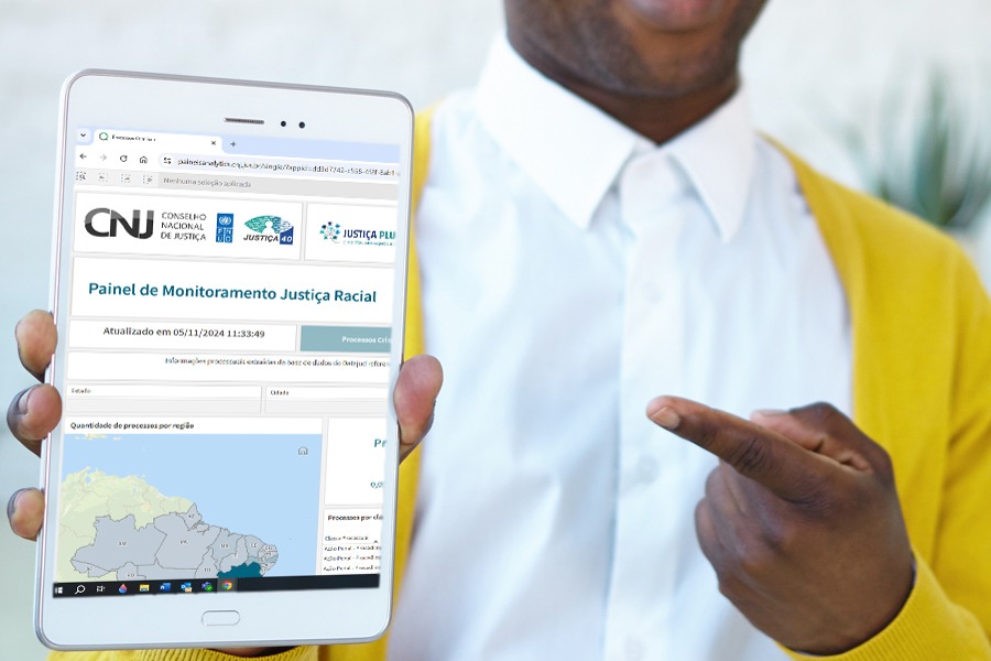 Uma pessoa negra, que veste uma camisa branca e um casaco amarelo, segura um tablet que apresenta a imagem da aba de um site, com a inscrição “Painel de Monitoramento Justiça Racial”, além de outras informações como logotipo do Conselho Nacional de Justiça (CNJ) na parede, juntamente com a inscrição “Conselho Nacional de Justiça”, e um mapa do Brasil. 