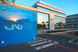 Fotografia da entrada do Conselho Nacional de Justiça (CNJ). Uma placa azul com a inscrição “CNJ Conselho Nacional de Justiça” aparece em destaque no lado esquerdo da foto. No lado direito, um pouco desfocado, o prédio espelhado do CNJ e na parede, o logotipo e a inscrição com o nome do órgão. Na parte inferior da foto, há uma faixa de pedestres além de uma linha vermelha pintada no chão. 