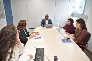 Fotografia de cinco pessoas em uma sala fechada. Elas estão sentadas ao redor de uma mesa e conversam. Há quatro mulheres e um homem. Em cima da mesa há alguns papéis, um caderno azul, garrafa de água, um teclado de computador e um notebook. 