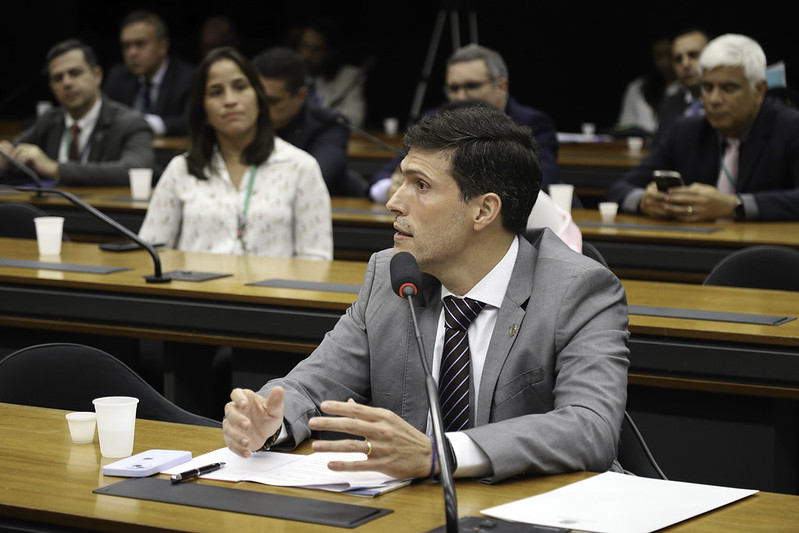 Fotografia do conselheiro do CNJ, Pablo Coutinho Barreto (homem branco, de cabelos escuros, veste camisa social branca, terno cinza e gravata vermelha) enquanto fala em um microfone. Ele está sentado atrás de uma mesa de madeira, que possui outros itens em cima, como um copo de água, um celular, alguns papéis e uma caneta. Ao fundo algumas pessoas o observam. Elas também estão sentadas atrás de mesas de madeira com microfones.
