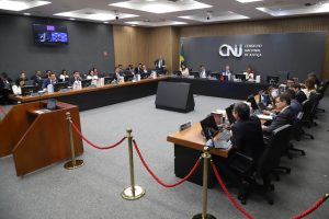 Fotografia do Plenário do CNJ, onde estão presentes o presidente do Conselho Nacional de Justiça (CNJ) e do Supremo Tribunal Federal (STF), ministro Luís Roberto Barros, e seus conselheiros. No fundo da foto, um painel cinza com o logotipo “CNJ” e a inscrição “Conselho Nacional de Justiça”. Há também um mastro com a bandeira do Brasil. No lado esquerdo da foto, uma televisão transmite a imagem do ministro e de uma intérprete de Libras. Todos estão sentados atras de uma mesa de madeira que está posicionada em formato de “U”. Em frente à mesa, há um cordão vermelho e um púlpito de madeira. 