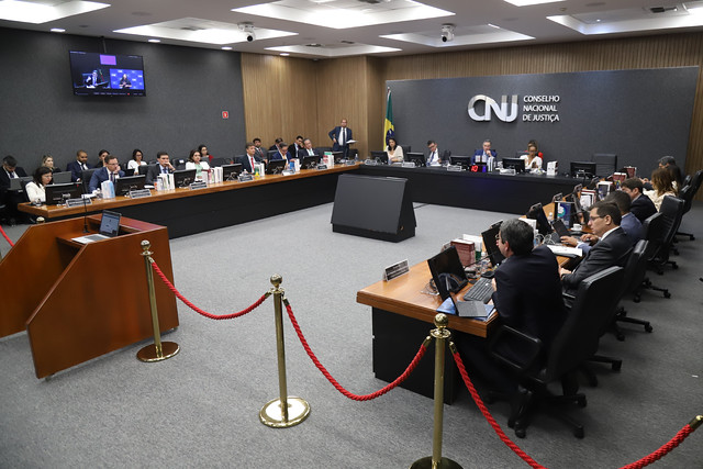 Fotografia do Plenário do CNJ, onde estão presentes o presidente do Conselho Nacional de Justiça (CNJ) e do Supremo Tribunal Federal (STF), ministro Luís Roberto Barros, e seus conselheiros. No fundo da foto, um painel cinza com o logotipo “CNJ” e a inscrição “Conselho Nacional de Justiça”. Há também um mastro com a bandeira do Brasil. No lado esquerdo da foto, uma televisão transmite a imagem do ministro e de uma intérprete de Libras. Todos estão sentados atras de uma mesa de madeira que está posicionada em formato de “U”. Em frente à mesa, há um cordão vermelho e um púlpito de madeira. 
