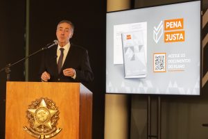 Fotografia do presidente do Conselho Nacional de Justiça (CNJ) e do Supremo Tribunal Federal (STF), ministro Luís Roberto Barroso (homem branco de cabelos grisalhos, veste uma camisa social branca, terno peto e gravata cor de vinho) enquanto fala em um microfone que está sobre um púlpito de madeira com o brasão do Brasil. Ao lado dele, uma televisão transmite a imagem de um card horizontal de cor cinza, e que possui a inscrição “PENA JUSTA” dentro de duas caixas de texto de cor laranja.  Embaixo, há uma segunda inscrição, “Acesse os documentos do plano” e ao lado há um QR Code. No lado esquerdo do card, a imagem de um livro de cor cinza. 