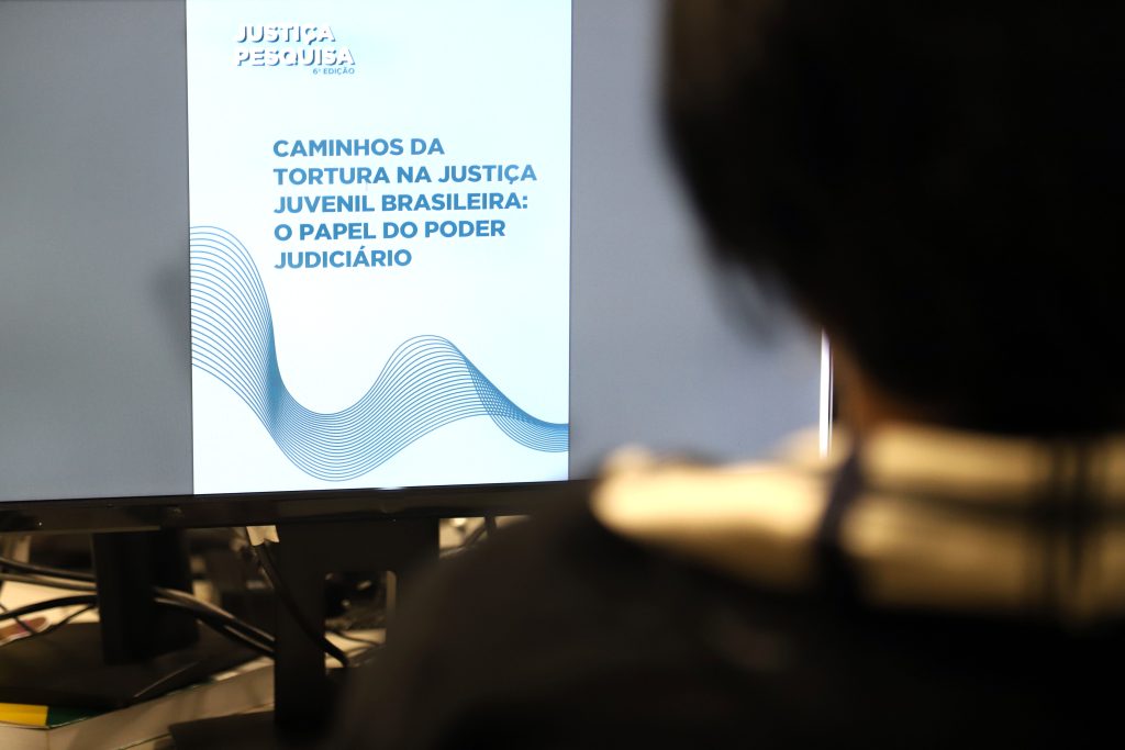 Fotografia de uma pessoa enquanto olha a tela de um computador que transmite a imagem de um card de fundo azul claro e possui a inscrição “Justiça Pesquisa” e “Caminhos da Tortura na Justiça Juvenil Brasileira: O Papel do Poder Judiciário”.    