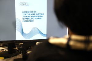 Fotografia de uma pessoa enquanto olha a tela de um computador que transmite a imagem de um card de fundo azul claro e possui a inscrição “Justiça Pesquisa” e “Caminhos da Tortura na Justiça Juvenil Brasileira: O Papel do Poder Judiciário”.    