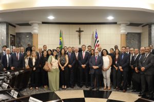Fotografia de diversas pessoas, entre eles o ministro Mauro Campbell, em frente a dois mastros com bandeiras do Brasil e do Maranhão e de um crucifixo que está na parede ao fundo. Eles estão no Tribunal de Justiça do Maranhão (TJMA). 