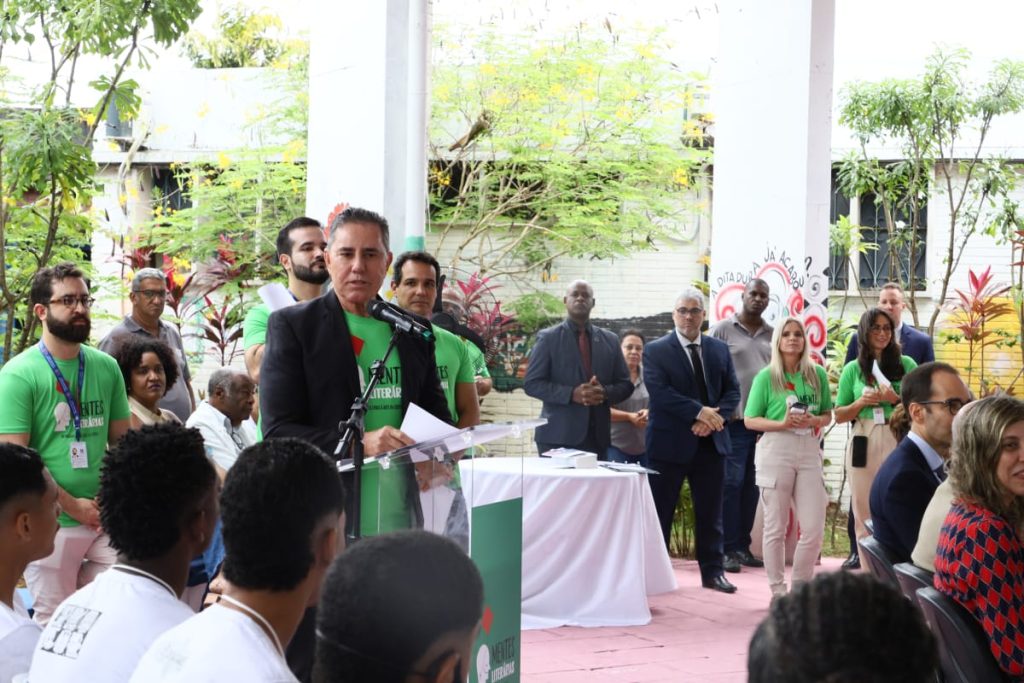 Homem fala ao microfone em evento ao ar livre; pessoas de camiseta verde e público sentado acompanham, com árvores ao fundo.