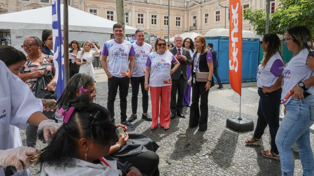 Pessoas em evento ao ar livre com tenda e banner do Senac; grupo observa corte de cabelo feito por profissionais em duas mulheres.