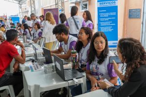  Pessoas sentadas em mesas com notebooks atendem público em evento social; banner azul indica serviços socioassistenciais.