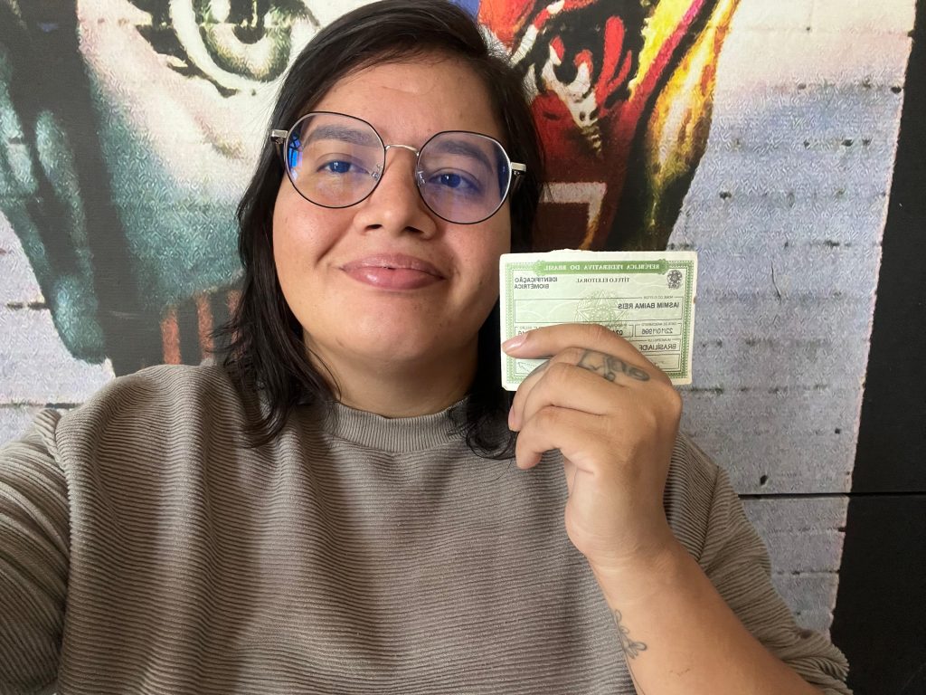 Pessoa de óculos segura um título de eleitor brasileiro na frente de um mural colorido, olhando para a câmera e sorrindo.