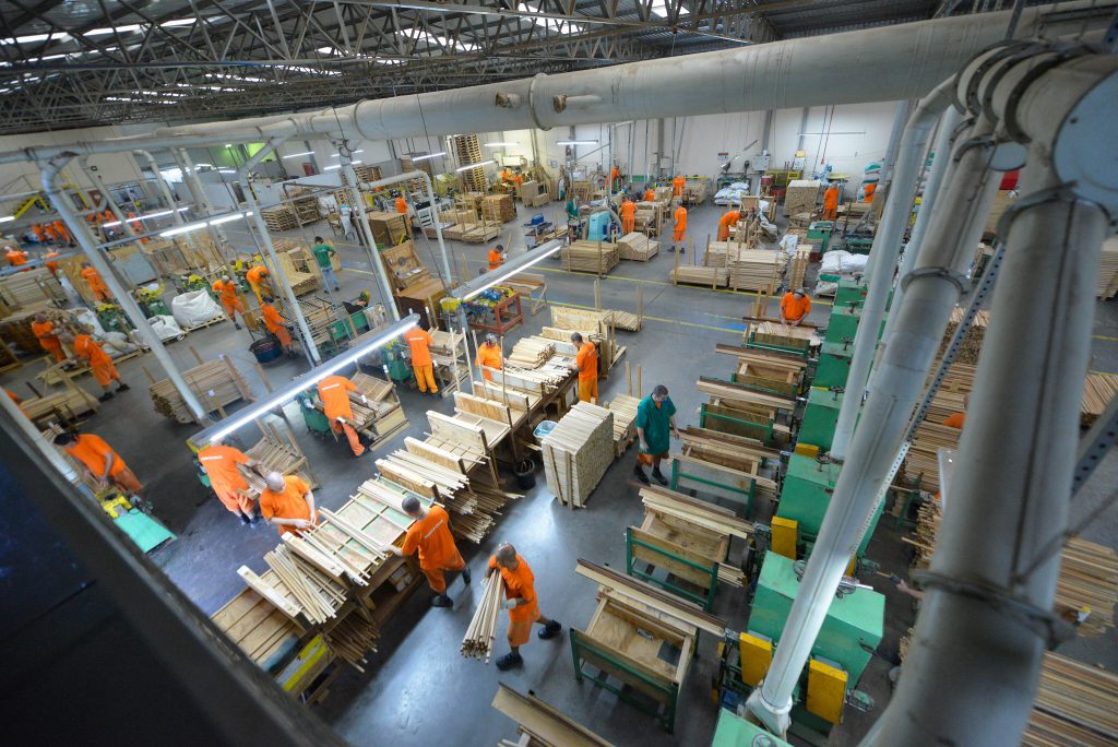 Fotografia. Mostra galpão industrial com pessoas em uniformes laranja e verde manuseando madeira em máquinas e bancadas.