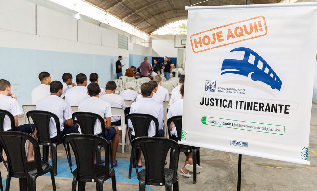 Fotografia. Jovens de costas, sentados em cadeiras, observam uma atividade. Em primeiro plano, um banner de divulgação com o texto "Justiça Itinerante".