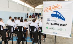 Fotografia. Jovens de costas, sentados em cadeiras, observam uma atividade. Em primeiro plano, um banner de divulgação com o texto "Justiça Itinerante".